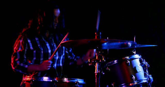 A young Caucasian female musician is playing the drums passionately in a low-light setting, with copy space. Her dynamic performance is accentuated by the dramatic lighting, capturing the energy of live music.