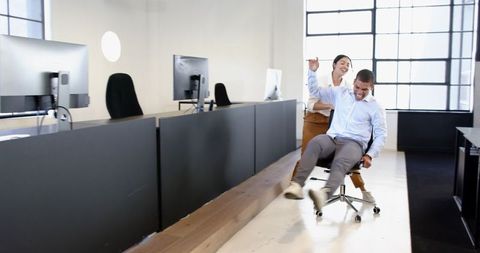 Colleagues playing cheerfully with office chair in modern workspace