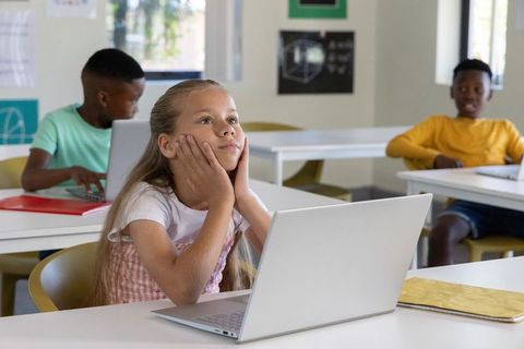 Schoolgirl daydreaming at laptop in modern classroom while classmates work quietly during class