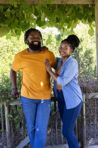 Sunlit african couple standing under grape pergola smiling and leaning on rustic railing