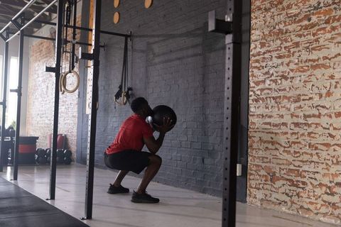 African american man squatting holding medicine ball wearing red shirt in industrial gym
