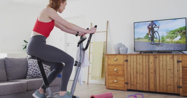 Caucasian woman watching tv and using elliptical trainer at home. Domestic life, healthy lifestyle and fitness, unaltered.