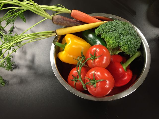 A collection of vibrant fresh vegetables including ripe tomatoes, yellow and red bell peppers, broccoli, and multicolored carrots placed in a stainless steel bowl. This photograph can be used for promoting healthy eating, organic food products, vegan and vegetarian lifestyle, and nutritional guides. Ideal for kitchen decor, dietary magazines, and online culinary blogs.