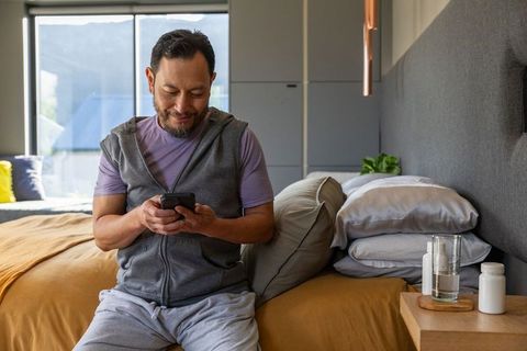 Mature asian man sitting on bed scrolling smartphone in modern bedroom morning light