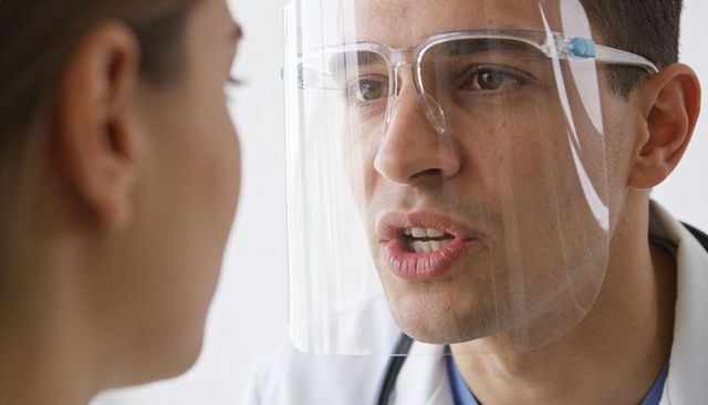 Male doctor wearing face shield communicating with patient during clinical consultation closeup