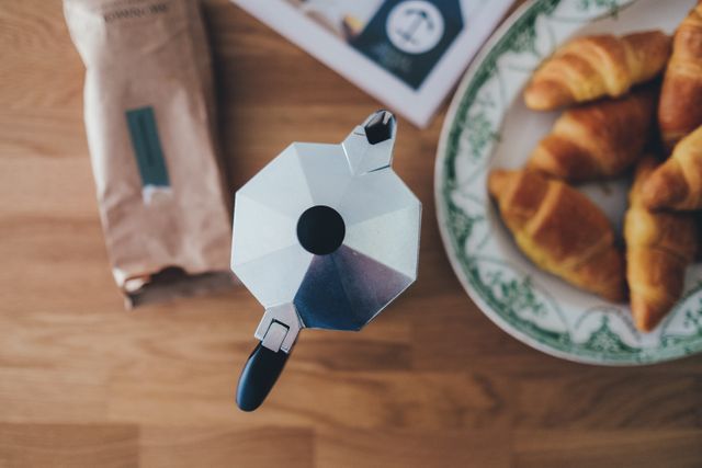 Aerial view of a wooden table featuring an Italian coffee maker next to four croissants on a decorative plate, alongside a bag of coffee. This image is great for websites or advertisements related to breakfast foods, kitchen decor, or coffee products. It can also be used in blogs or social media posts about morning routines or traditional breakfast recipes.