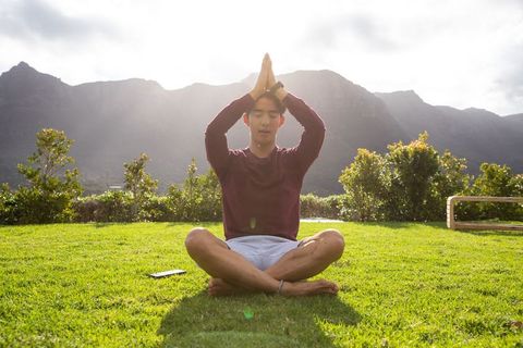 Asian man meditating cross-legged on grass with mountains and sunbeams at sunrise