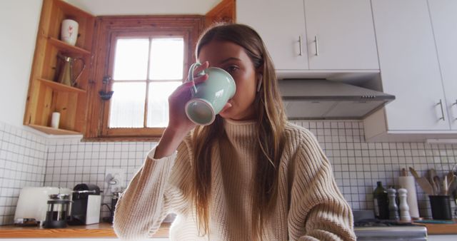 Young woman is drinking coffee in a cozy home kitchen. She is wearing a white sweater and enjoying a moment of relaxation during her morning routine. Perfect for illustrating lifestyle, home living, relaxation, and self-care themes.