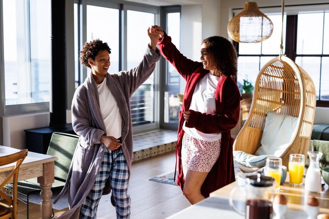 Happy, diverse lesbian couple having fun dancing at breakfast in sunny kitchen. Togetherness, free time and domestic life concept.