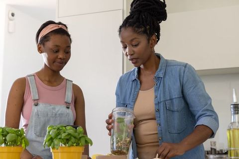 African american mother teaching teen daughter making green smoothie with fresh basil