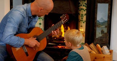 Father teaching son guitar by cozy fireplace