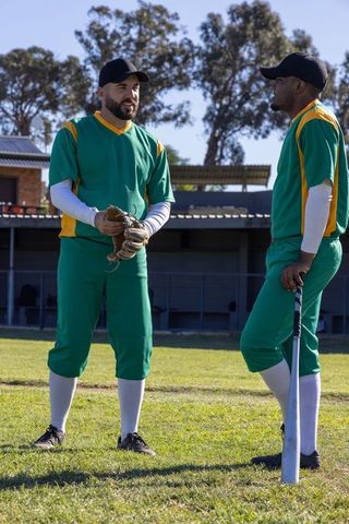 Discussing pregame strategy, two baseball teammates wearing green uniforms on grass field