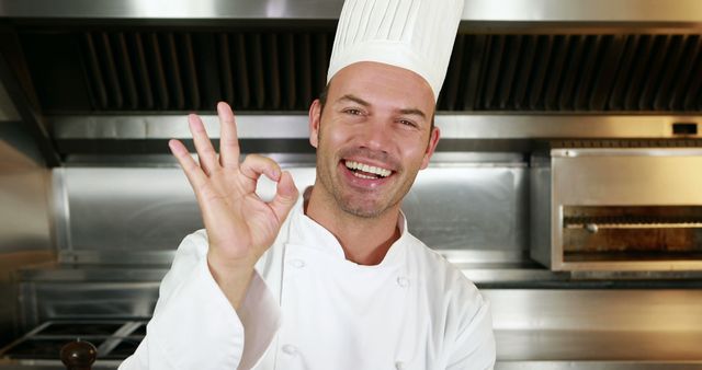 Chef in white uniform and hat smiling while showing OK gesture in professional kitchen. Perfect for illustrating concepts of culinary expertise, restaurant services, positive feedback, happiness in workplace, successful cooking, main cover for culinary blogs, advertisements for culinary schools, or restaurant promotions.