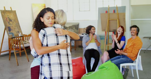 Two women are embracing in a supportive hug in an art studio, with copy space. Diverse individuals are seated in the background, engaging in cheerful conversation, creating a warm, communal atmosphere.