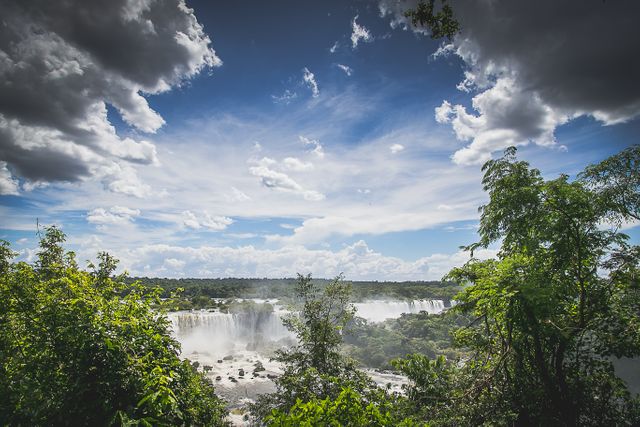 Breathtaking view of Iguazu Falls surrounded by lush green trees under vibrant sky with clouds. Ideal for travel blogs, tourism promotion, nature conservation campaigns, or landscape photography portfolios.