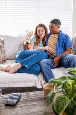 Young couple cuddling on sunlit sofa relaxing at home with mugs and cozy cushions