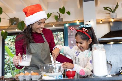 Diverse mother and daughter baking christmas cookies, mixing batter and smiling in kitchen