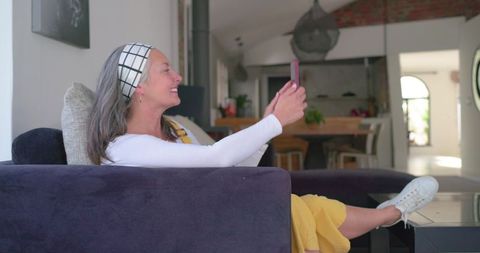 Senior woman making video call on smartphone while relaxing on purple sofa in open living room
