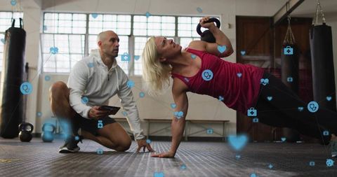 Woman performing kettlebell side plank while coach monitoring workout with tablet and icons