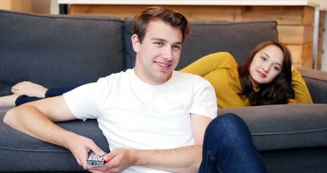 Young man holding TV remote while woman reclines on couch behind. They are in casual wear and appear relaxed and happy. This can be used for themes related to leisure, home life, couples spending time together, weekend activities, and relaxation at home.