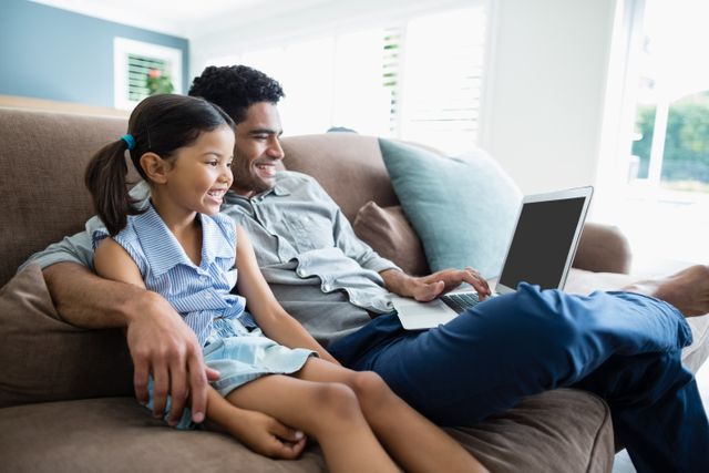 Father and daughter sitting on a sofa, using a laptop together. They are smiling and enjoying their time. Ideal for concepts related to family bonding, technology use at home, parenting, and leisure activities.