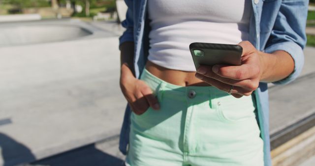 Young woman wearing denim jacket and mint green shorts engaged with her smartphone outdoors on a sunny day. Ideal for use in contexts such as modern technology, digital lifestyle, summer activities, casual fashion, and social media engagement.