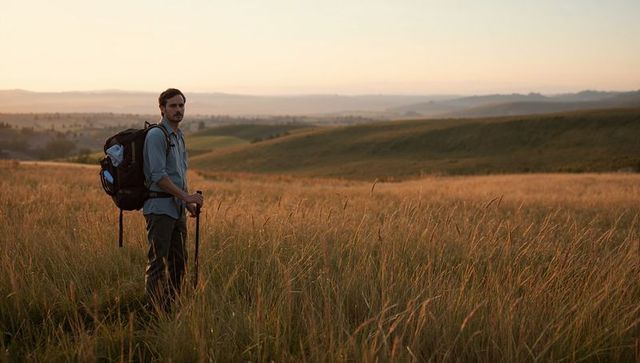 Male hiker standing in golden meadow at sunset holding trekking poles and backpack
