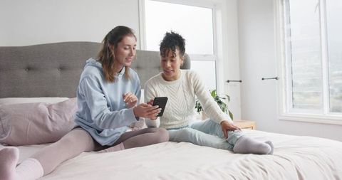 Multiracial friends relaxing on bed in cozy loungewear sharing smartphone moment