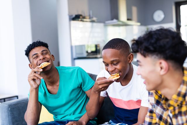 Happy diverse teenager male friends eating pizza and sitting on sofa at home. Hanging out with friends and spending quality time together concept.