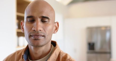 Serene bald man closing eyes resting in modern kitchen with stainless steel fridge