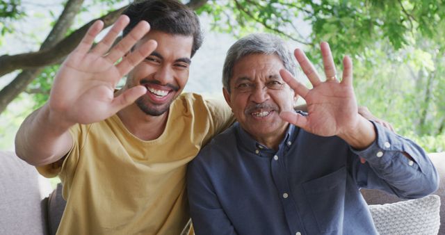 Two men, a young adult and an elder, are waving at the camera with big smiles while seated in an outdoor location. Their expressions convey joy and warmth, making it perfect for themes of family bonds, generational connection, happiness, and togetherness. Can be used in advertisements or articles that focus on family relationships, community connections, or outdoor activities.