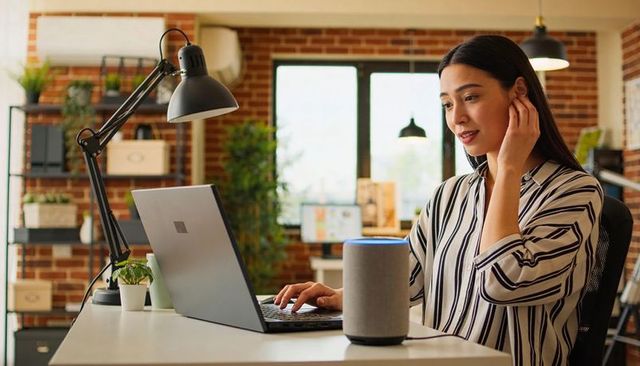Woman speaking to smart speaker while typing on laptop in home office with brick wall and plants