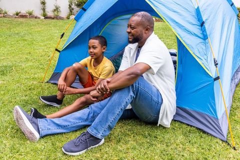African american father and son camping in backyard tent, relaxing on grass