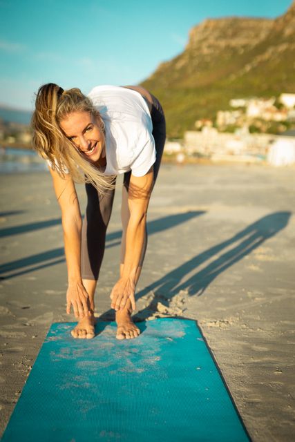 Woman stretching on a beach during sunset, smiling at the camera. Ideal for promoting healthy lifestyles, outdoor fitness, yoga retreats, and wellbeing programs. Perfect for use in fitness blogs, wellness websites, and advertisements for activewear or yoga equipment.