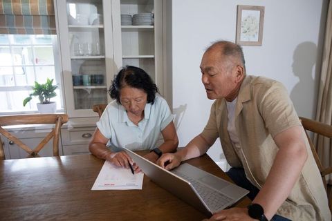Senior asian couple reviewing finances with laptop and paperwork at home kitchen table