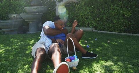 African american dad playing and laughing with teenage son on sunny backyard lawn with easter basket