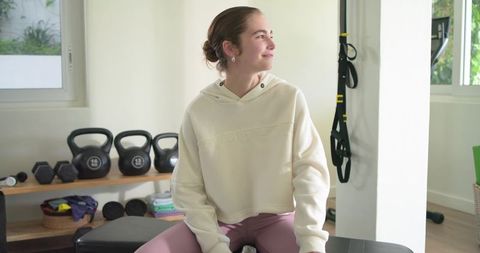 Smiling woman sitting on bench in sunlit home gym wearing cream hoodie and kettlebells