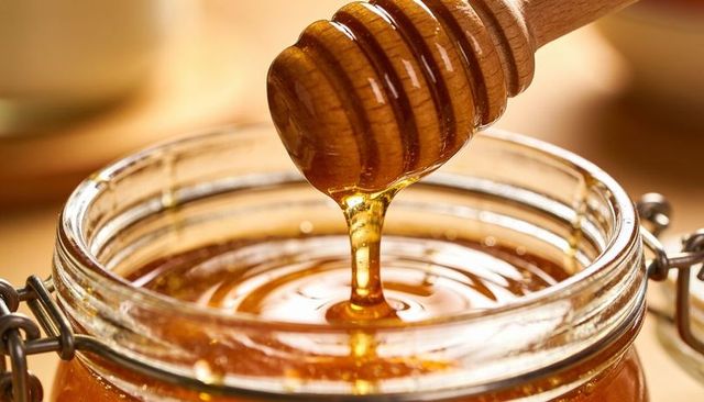 Golden honey dripping from wooden dipper into glass jar creating glossy amber ripples
