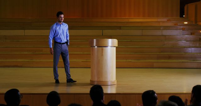 Man standing on stage in front of wooden podium in auditorium, engaging audience. Perfect for illustrating public speaking, presentations, educational events, seminars, conferences, and business meetings.