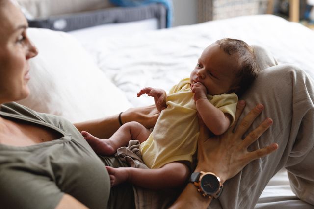 Caucasian mid adult mother holding her newborn baby while sitting on a bed at home. The baby is wearing a yellow onesie and appears content. This image is perfect for use in parenting blogs, family-oriented advertisements, or articles about motherhood and newborn care.