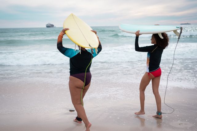Two diverse female friends are seen from the back, carrying surfboards as they walk towards the ocean. The scene captures the essence of summer, adventure, and friendship. Ideal for use in travel promotions, lifestyle blogs, and advertisements focusing on outdoor activities, sports, and beach vacations.