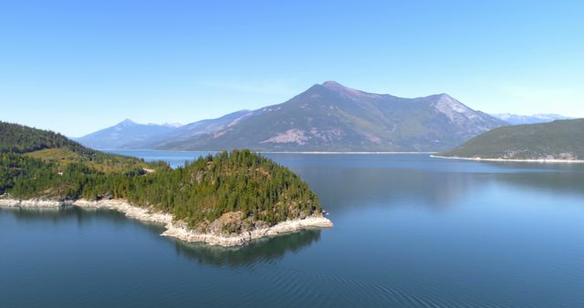 A scenic drone view captures a serene mountain lake surrounded by forested hills under a clear blue sky. The calm water reflects the natural beauty of the landscape, contributing to a tranquil atmosphere. Ideal for promotional material related to travel, nature conservation, outdoor activities, and environmental campaigns.