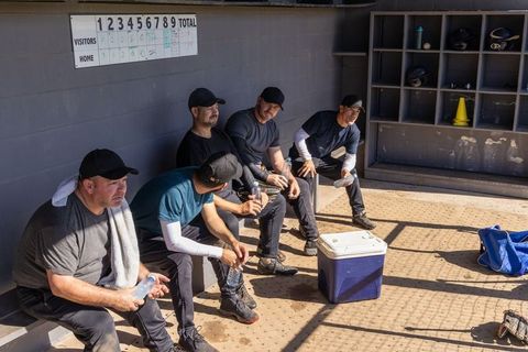 Diverse baseball teammates resting in dugout drinking water under sunlit shadows