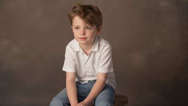 Thoughtful young boy sitting on wooden stool wearing white shirt and blue jeans studio portrait