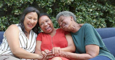 Three diverse mature women laughing and holding hands on outdoor sofa in garden