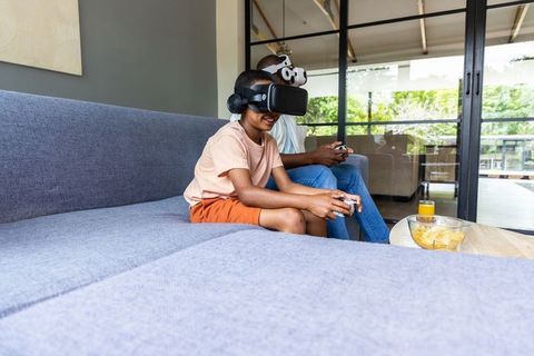 African american father and teen son playing vr games on gray sofa with controllers