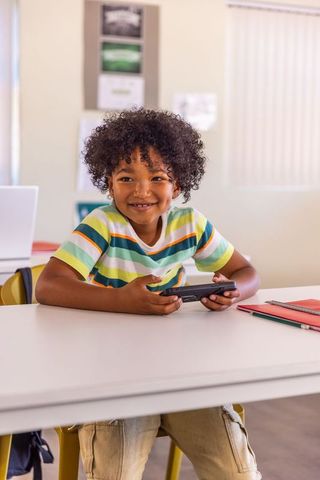 Smiling african american schoolchild holding handheld device while learning in classroom