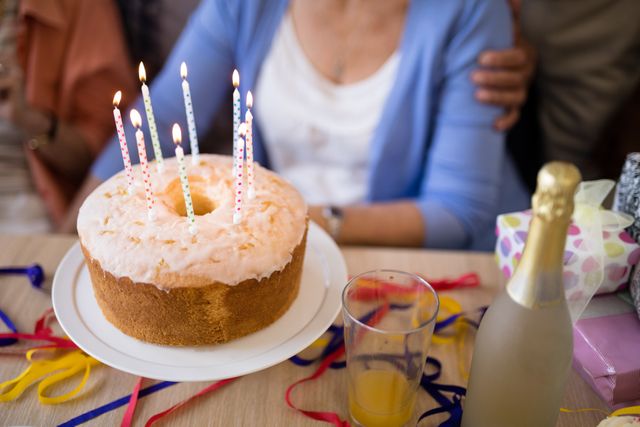 This image captures a close-up of a birthday cake with lit candles, surrounded by senior individuals celebrating. The scene includes festive elements like colorful ribbons, a glass of juice, and a bottle of champagne, suggesting a joyful and special occasion. Ideal for use in birthday celebration promotions, senior community events, and festive greeting cards.