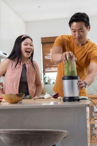 Asian couple preparing green smoothie on kitchen island blending vegetables together