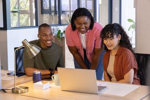 Diverse team collaborating around laptop, smiling and leaning over modern open-plan desk